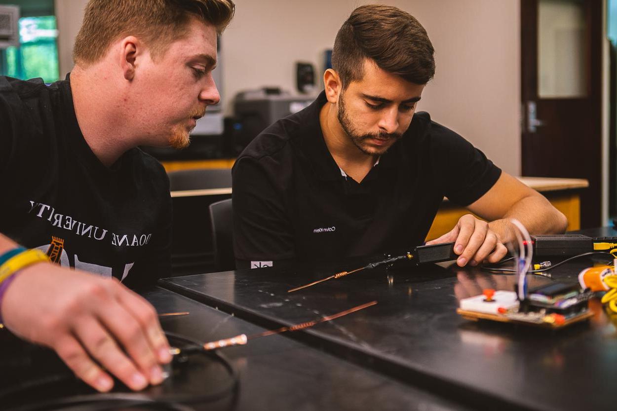 Junior Jeremey Allgeyer倾向于Henrique Henriques, senior (left), 查看一个黑色小装置上的读数，测量他手持的发射器的无线电信号.